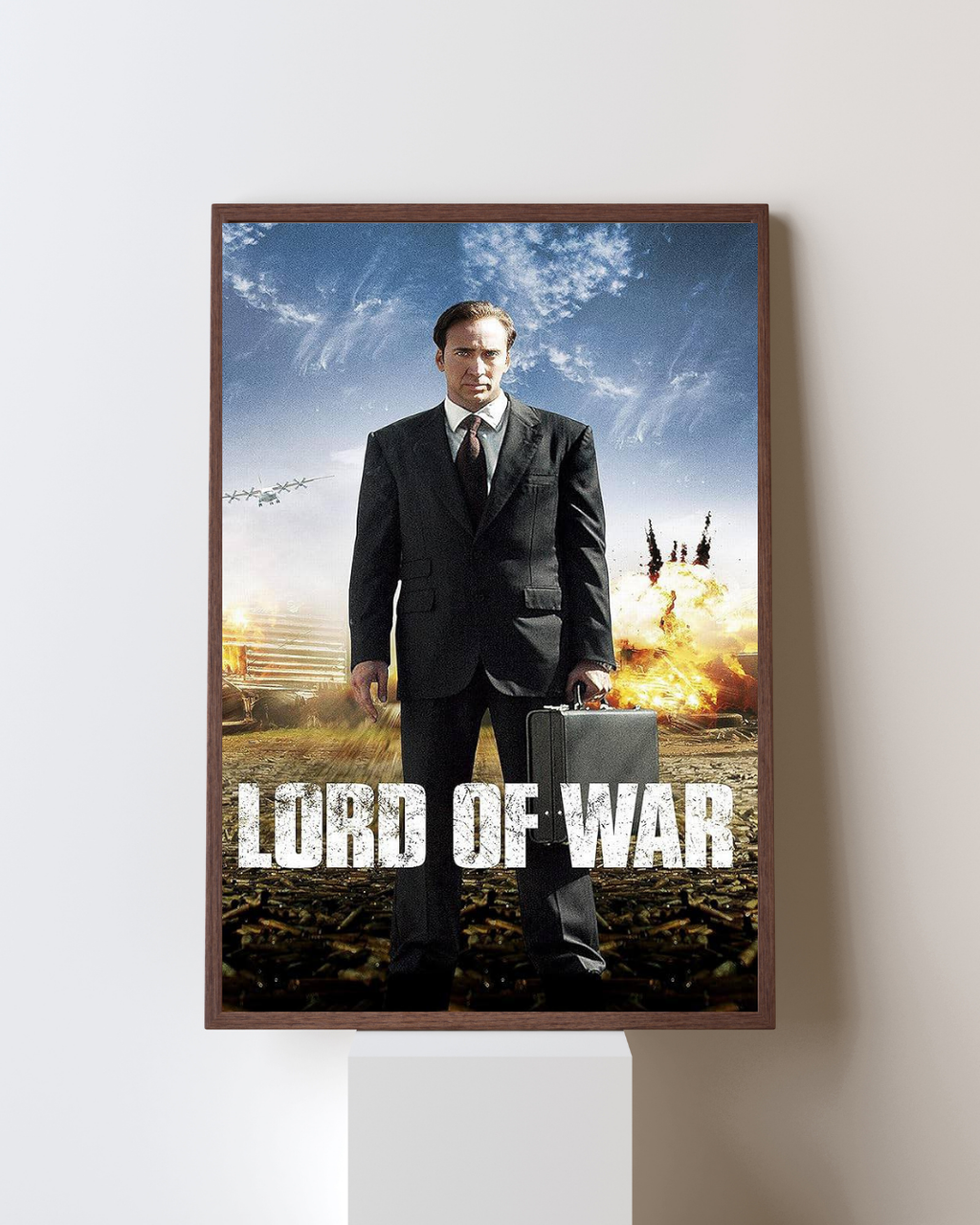 Frame Lord of War movie poster featuring Nicolas Cage standing in a suit holding a briefcase on a battlefield covered with bullet casings and explosions in the background, printed in high definition with Canon HD inks.